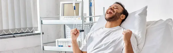 Jeune patient indien heureux acclamant et gesticulant activement tandis que dans le lit d'hôpital, soins de santé, bannière — Photo de stock