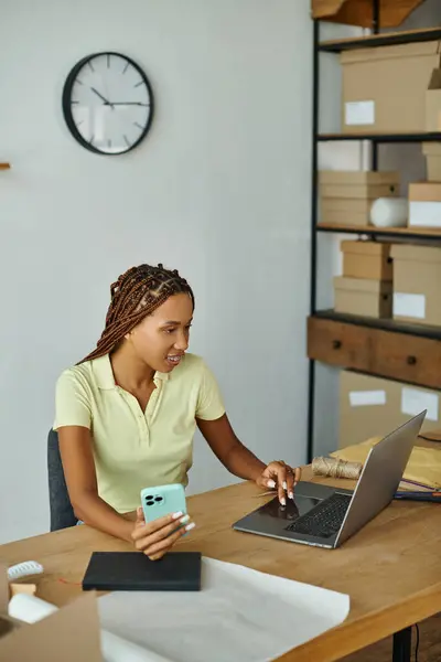 Fröhlich attraktive afrikanisch-amerikanische Verkäuferin prüft etwas auf ihrem Laptop und hält ihr Smartphone in der Hand — Stockfoto