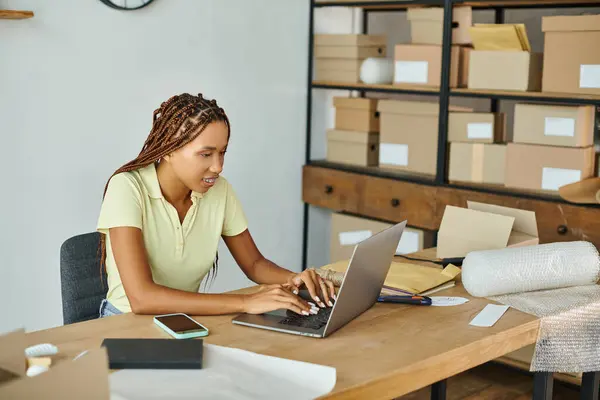 Joyeux vendeur afro-américain en tenue décontractée travaillant dur sur son ordinateur portable avec des colis sur toile de fond — Photo de stock