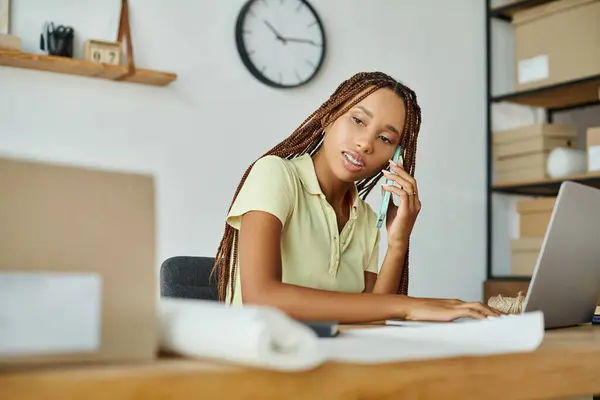 Atractiva mujer afroamericana comerciante hablando por teléfono mientras trabaja en sus paquetes, entrega - foto de stock