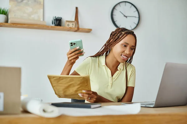 Joyeux jeune afro-américaine femme commerçante tenant téléphone et paquet postal et regardant son ordinateur portable — Photo de stock