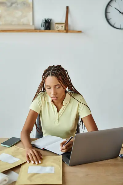 Attractive african american female seller taking notes and looking at post packet, delivery concept — Stock Photo