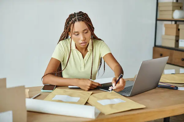 Atractiva mujer afroamericana comerciante tomando notas mientras trabaja duro, concepto de entrega - foto de stock
