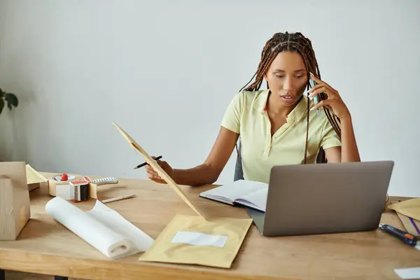 Young african american female merchant talking by phone holding post packet and looking at laptop — Stock Photo