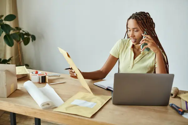 Весела приваблива афроамериканська жінка-продавець розмовляє по телефону і дивиться на поштовий пакет — стокове фото