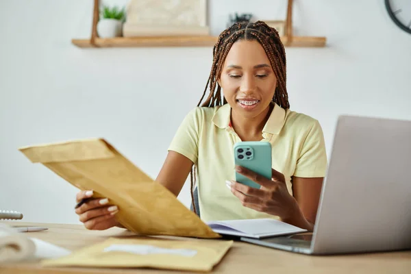 Joyeux détaillant afro-américain tenant un paquet de courrier et regardant son smartphone, la livraison — Photo de stock