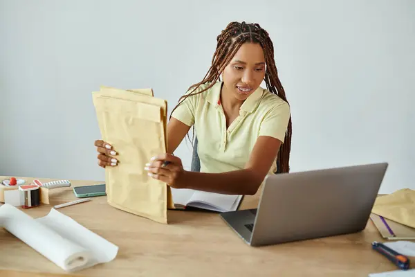 Радостная африканская торговая женщина держит почтовые пакеты и смотрит на ноутбук, доставку — стоковое фото