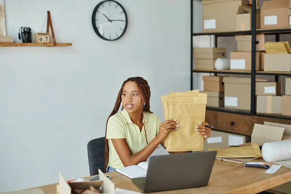 Jolly attractive african american female seller holding post packets and looking away, delivery — Stock Photo