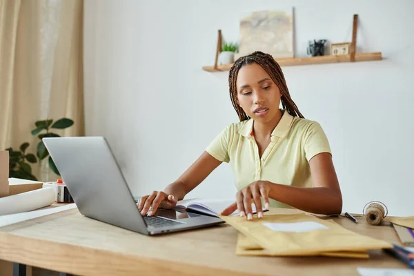 Atraente Africano americano varejista feminino trabalhando no laptop e olhando para pacote post atentamente — Fotografia de Stock