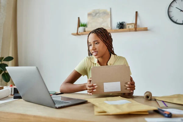 Belle vendeuse afro-américaine avec bretelles emballage boîte en carton et en regardant son ordinateur portable — Photo de stock