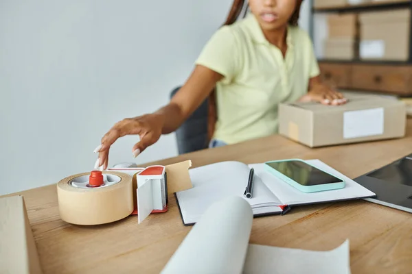 Cropped view of young african american female seller using tape to pack cardboard box, delivery — Stock Photo