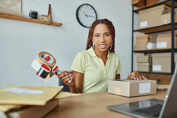 Attractive jolly african american female retailer using tape on cardboard box and smiling at camera — Stock Photo