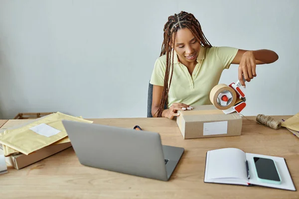 Joyeux jolie afro-américaine femme en utilisant la bande sur la boîte tout en travaillant dur, concept de livraison — Photo de stock