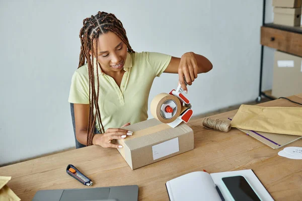 Jolly young african american female retailer using tape on cardboard box, delivery concept — Stock Photo