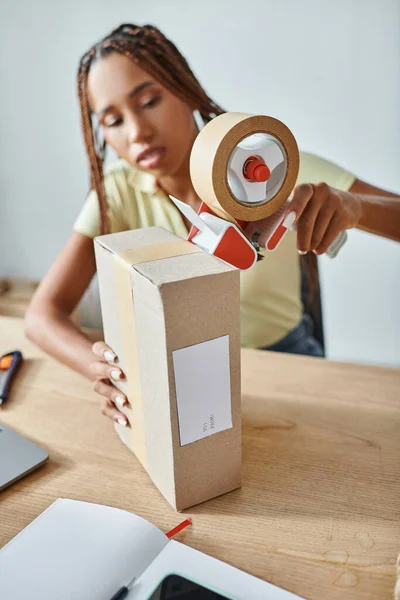 Concentrarsi sulla scatola di cartone con donna afroamericana offuscata utilizzando nastro adesivo su di esso, concetto di consegna — Foto stock