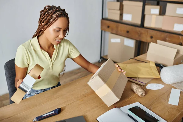 Alegre jovem africano americano vendedor feminino segurando fita e olhando para caixa de papelão, entrega — Fotografia de Stock