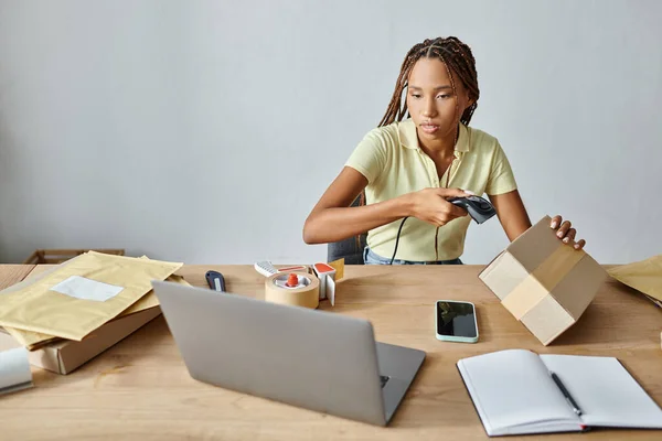 Beautiful african american female merchant scanning box and looking at laptop, delivery concept — Stock Photo