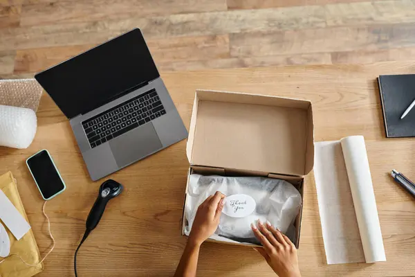 Cropped view of hard working african american female retailer packing box next to laptop, delivery — Stock Photo