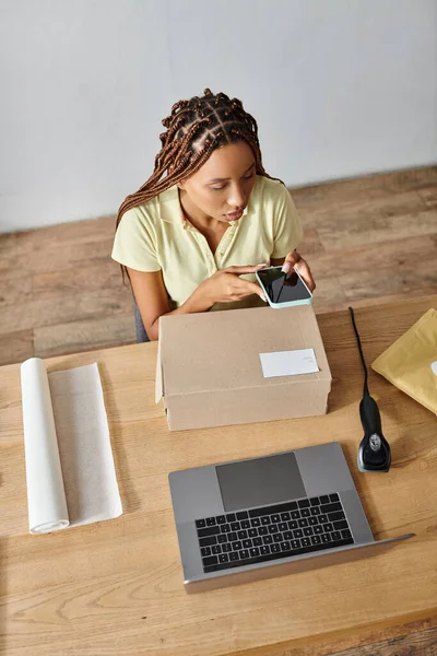 Gut aussehende junge afrikanisch-amerikanische Verkäuferin schaut auf ihr Handy neben Laptop und Schachtel — Stockfoto