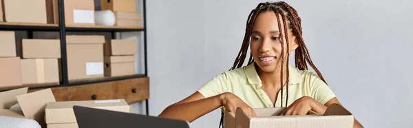 Joyous attractive african american female seller packing box and looking at laptop, delivery, banner — Stock Photo