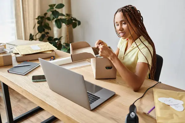 Jolly young african american female seller packing box and looking at laptop, delivery concept — Stock Photo