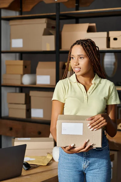 Bien parecido alegre africano americano mujer comerciante sosteniendo caja de cartón y mirando hacia otro lado, entrega - foto de stock
