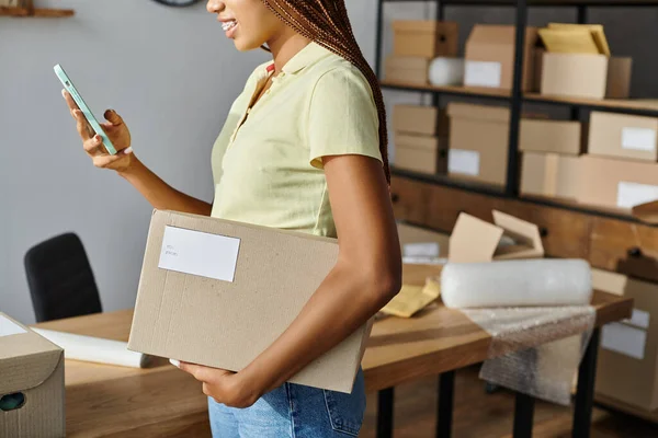 Cropped view of young african american woman with braces holding box and phone during work, delivery — Stock Photo