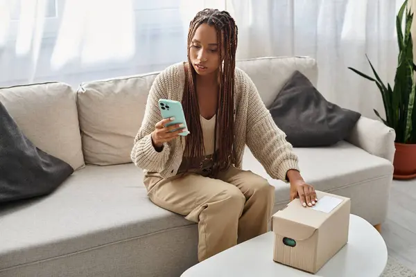 Séduisante jeune femme afro-américaine regardant son smartphone avant d'ouvrir le colis en carton — Photo de stock