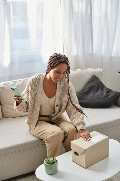 Cheerful attractive african american woman holding phone and preparing to open cardboard box — Stock Photo