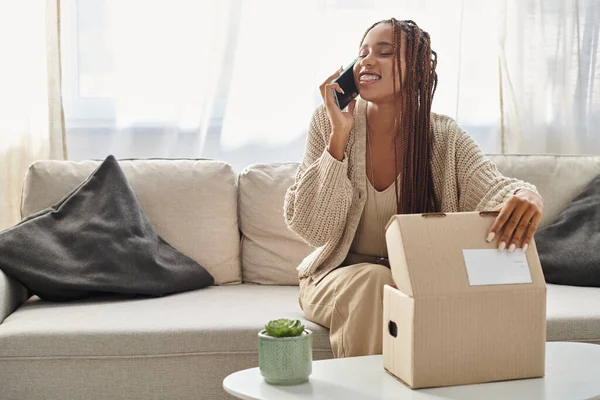 Joyeux attrayant afro-américain femme dans confortable homewear parler par téléphone avec la main sur le colis — Photo de stock