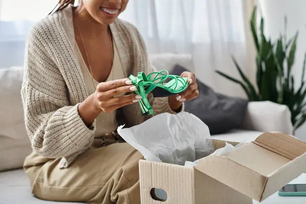 Vista ritagliata di gioiosa donna africana americana con bretelle disimballaggio le sue nuove scarpe verdi a casa — Foto stock
