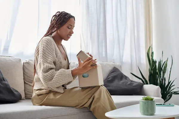 Joyeuse femme afro-américaine en tenue de maison avec bretelles regardant son colis assis sur le canapé — Photo de stock