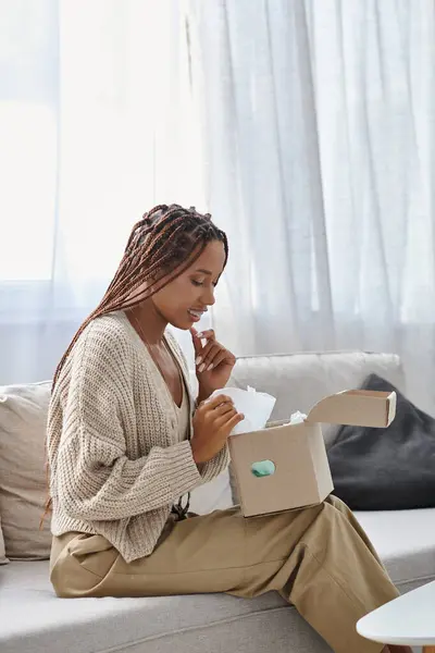 Belle femme afro-américaine joyeux assis sur le canapé et regardant son colis en carton — Photo de stock