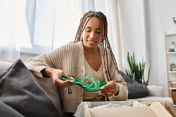 Alegre bien parecido africano americano mujer en la ropa de casa sentado y mirando sus nuevos zapatos verdes - foto de stock