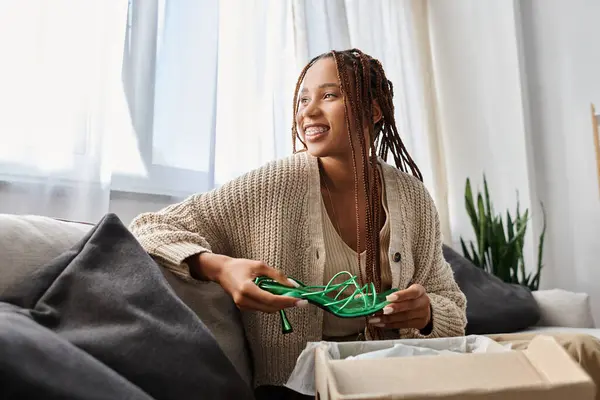 Attrayant joyeux afro-américain femme assise sur le canapé avec des chaussures vertes dans les mains et regardant loin — Photo de stock