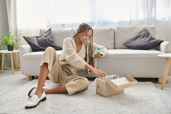 Joyeuse jeune femme afro-américaine en tenue de maison assise sur le sol avec téléphone et regardant le colis — Photo de stock