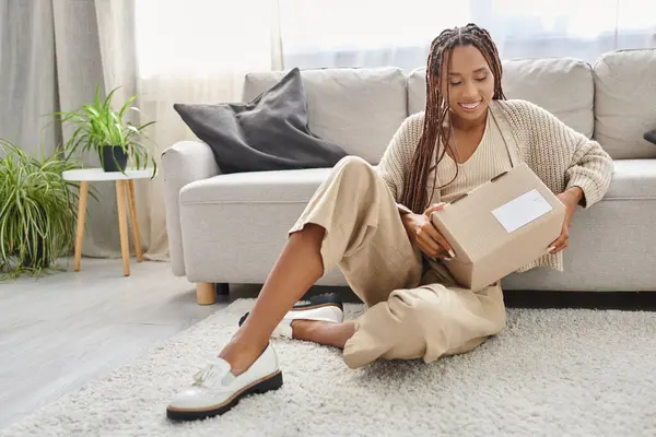 Joyeuse femme afro-américaine attrayante assise sur le sol et regardant son colis en carton — Photo de stock