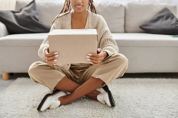 Vista recortada de la alegre mujer afroamericana con aparatos ortopédicos sentados en el suelo y sosteniendo el paquete - foto de stock