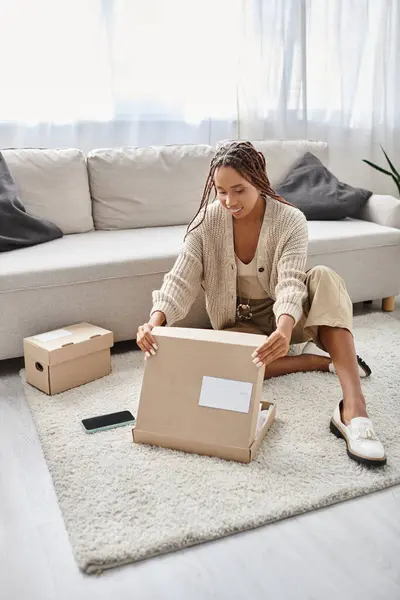 Joyous good looking african american woman in homewear sitting on floor and unboxing her parcel — Stock Photo