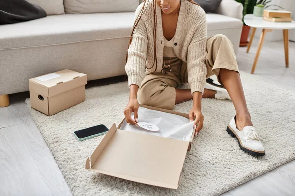Cropped view of jolly young african american woman sitting on floor and opening cardboard parcel — Stock Photo