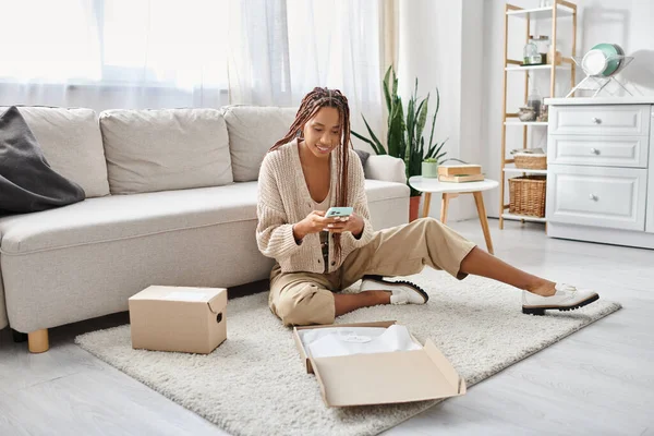 Heureux attrayante femme afro-américaine en vêtements de maison assis sur le sol et en regardant son téléphone — Photo de stock