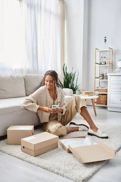 Alegre atractivo afroamericano mujer en ropa de casa sosteniendo el teléfono y mirando paquetes en el suelo - foto de stock