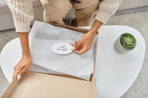 Cropped view of african american woman in cozy attire opening parcel with thank you sticker on it — Stock Photo