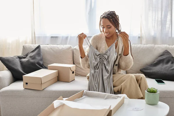 Femme afro-américaine gaie en vêtements de maison déballer sa nouvelle robe d'argent tout en étant assis sur le canapé — Photo de stock