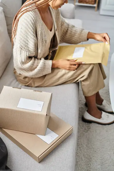Cropped view of african american woman with braces opening post packet while sitting on sofa — Stock Photo