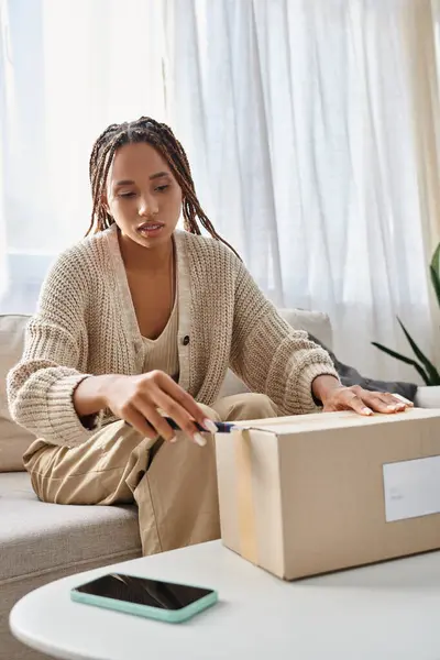 Young attractive african american woman in homewear using stationery knife to open her parcel — Stock Photo