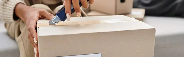 Cropped view of african american woman using stationery knife to open cardboard parcel, banner — Stock Photo