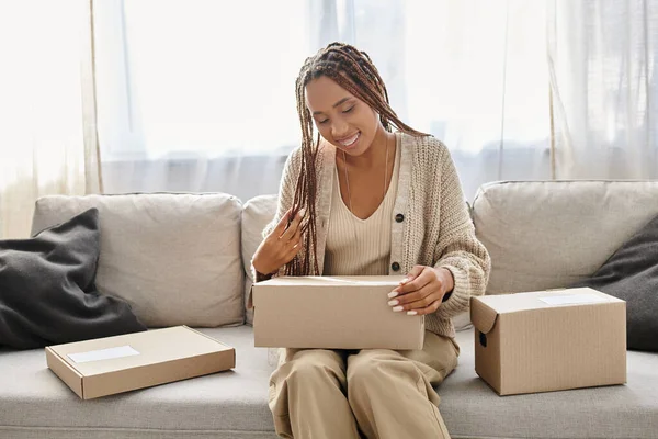 Belle femme afro-américaine joyeuse en tenue de maison assise sur le canapé et regardant la boîte en carton — Photo de stock