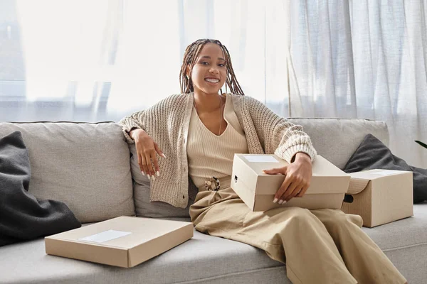 Joyeuse jeune femme afro-américaine en tenue confortable assise sur un canapé avec des boîtes et souriant à la caméra — Photo de stock