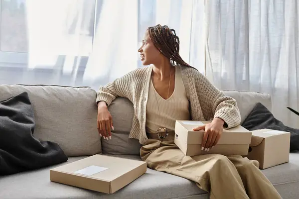 Cheerful african american woman in homewear sitting on sofa with cardboard boxes and looking away — Stock Photo